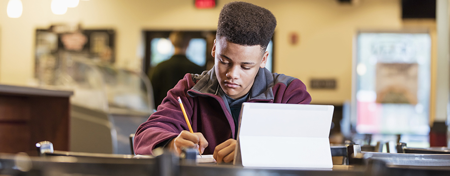 A man sits at a table and writes with a pencil. He is wearing a maroon sweater and has his laptop open in front of him.