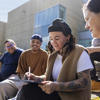 Anisa Francoeur sits outdoors with classmates on a sunny day and draws using her tablet.