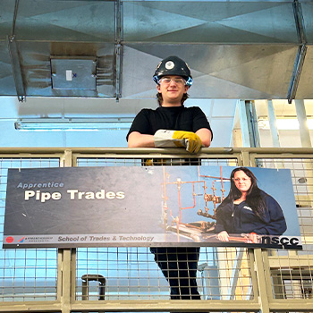 Austin standing while leaning over a rail in front of a Pipe Trades sign