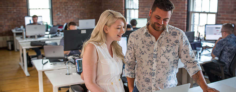 A blond woman and a bearded man stand shoulder to shoulder at a desk and look at an Apple laptop. There is a brick-walled office space in the background with several desks and people.