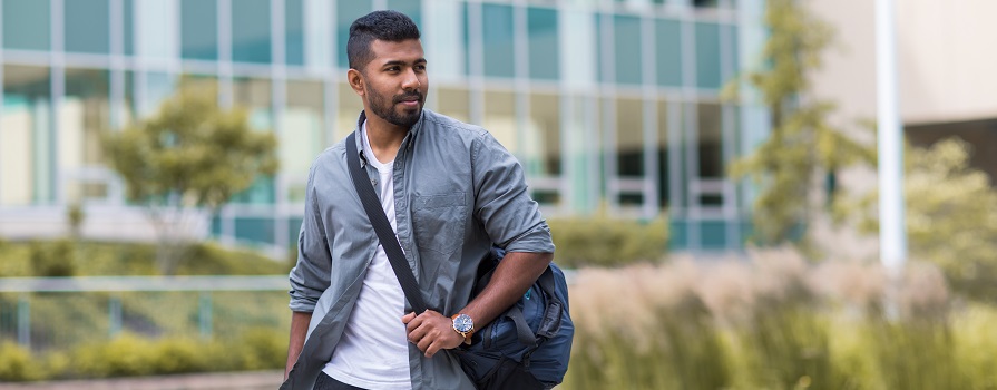 A man in an open button up shirt and t-shirt walks in front of an NSCC campus building wearing a crossbody bag.