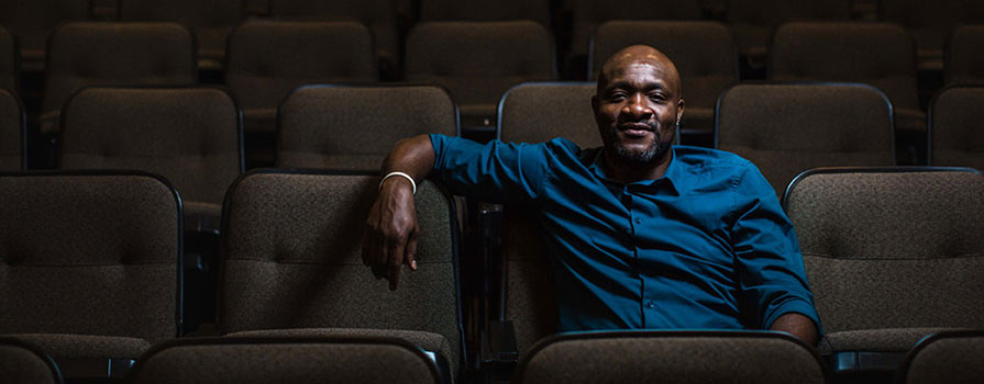 A man sits in an empty row of an auditorium. He is wearing a blue, button up shirt and looks at the camera with a smirk.