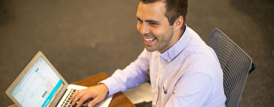 A man sits in a grey chair at a brown desk and types. He is smiling broadly and looking off to the left. He is wearing a light pink button-up shirt.