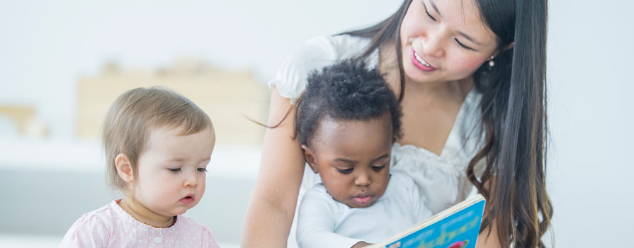 A woman reads to two very young children. One is sitting on her lap.