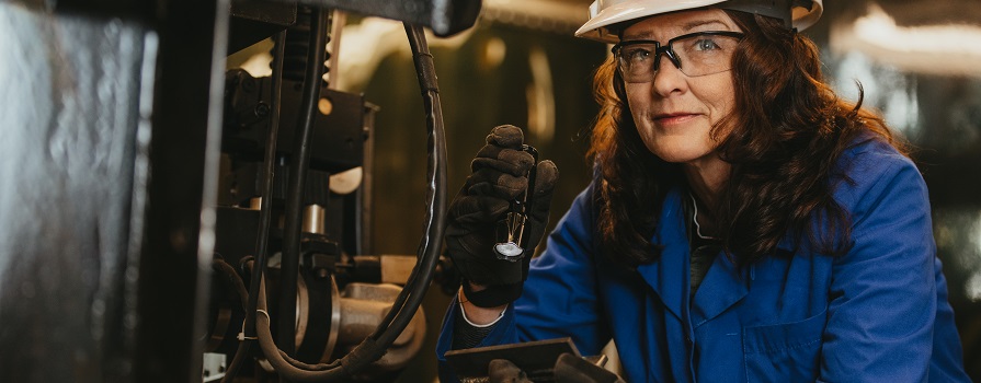 Christine MacKenzie inspects the weld on a piece of equipment with a flashlight. She is wearing a white hardhat, a blue smock, safety glasses and black gloves.