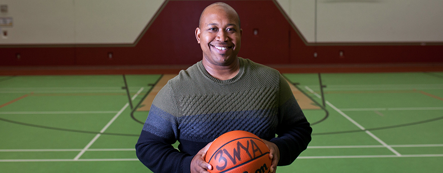 A man holding a basketball and wearing a sweater smiles at the camera. The ball is signed. He is in a basketball court with green flooring.