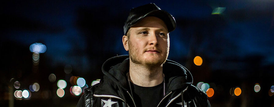 A young man wearing white pants, a black and white coat, black t-shirt and black hat, stands on a sidewalk and looks directly at the camera. It is night and the streetlights are illuminated. 