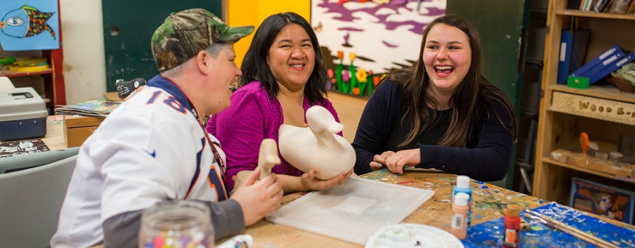 Two women and a man sit around a table. They are laughing and there are crafting supplies in front of them.