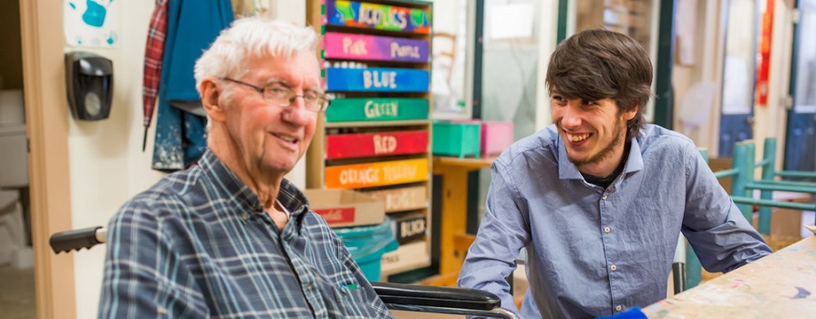 Two men sit at a table. One man is smiling at the other, who is in a wheelchair. They are in a classroom environment.