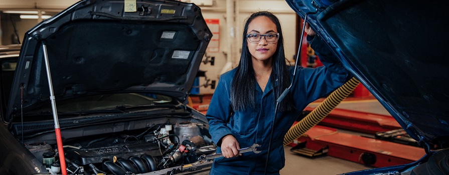 A young woman wearing blue coveralls stands in front of two cars with open engine bays. She is smiling and holds a wrench. She is in an automotive shop.
