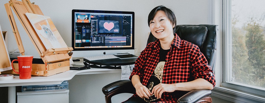 A woman wearing a plaid shirt sits at a computer. She is looking at the camera and her back is to her screen. She is holding a pen in her right hand.