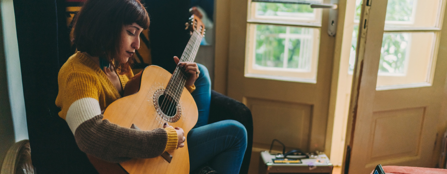 A woman in a yellow shirt strums a brown, acoustic guitar. There are two, open French doors seen in the background.
