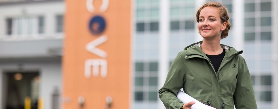 A woman in a green, zip-up jacket holds a rolled up map under her arm and walks in front of a building with the letters C O V E on it. 