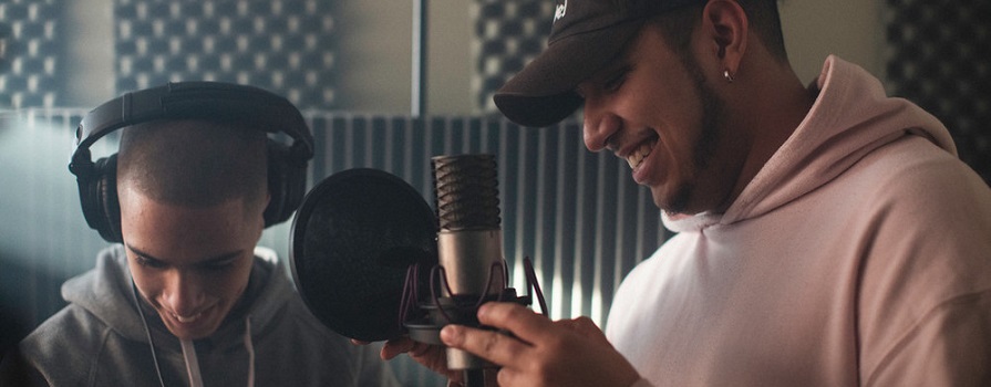 Two men smile while standing close to a professional-looking microphone. They are in a sound booth.