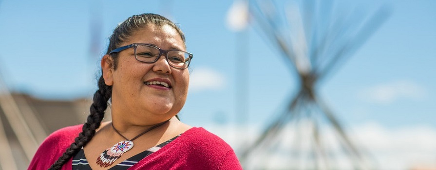 A woman in glasses smiles while looking off to the right of the image. She is wearing a maroon, button up sweater, a black and white striped t-shirt and a beaded necklace. He hair is in a braid. In the background, two teepee frames can be seen. 