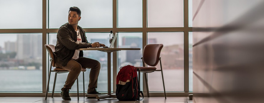 A man sits at a table and works on a laptop in front of a wall of windows.