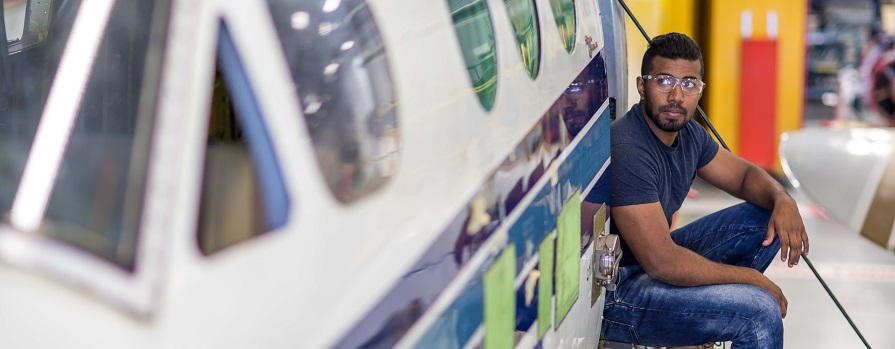 A man looks up while working underneath a plane. He is wearing blue work clothes and safety glasses. He is in a large, open workspace.