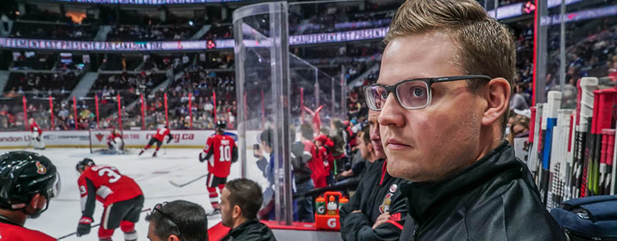 A man in glasses looks at something unseen to the left of the image. He is standing in the bench area of a large hockey stadium. People can be seen seated in front of him and playing hockey on the ice. There are many spectators in the background.