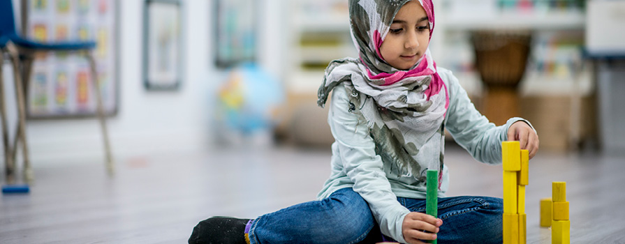 A young girl wearing a hijab sits on the floor playing with blocks.