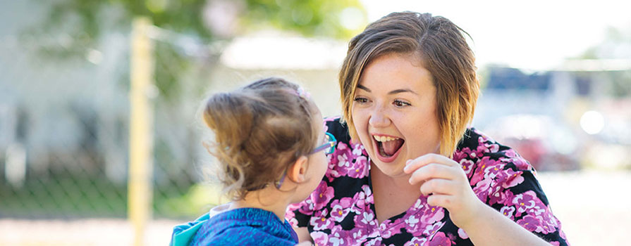 A young woman in a flowery, pink shirt smiles with an open mouth at a young child. They are seated at a picnic table in a park on a sunny day. 