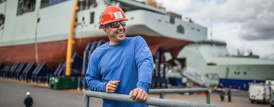 A man in a blue shirt, safety glasses and orange hardhat looks off to the top, right of the image. In the background there is a partially-constructed navy ship and a tall crane.