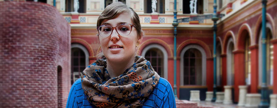 A woman wearing glasses, a blue sweater and scarf, stands in the atrium of a classically styled building with a glass domed roof.