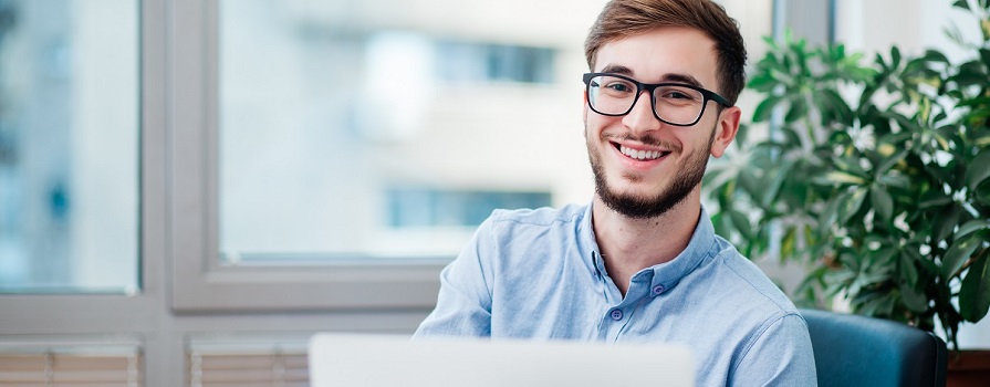 A man in glasses and a button up shirt smiles. He is in front of large windows and a plant. He appears to be working on a laptop.