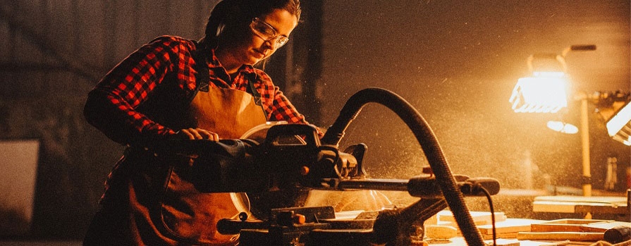 A woman in safety glasses and a plaid shirt works with a reciprocating saw.