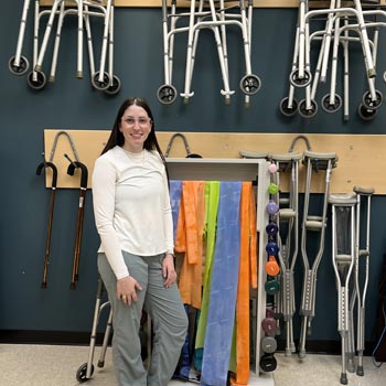 Jessie, an Occupational Therapy / Physiotherapy Assistant student at NSCC is standing in the program's lab with various OT/PT equipment behind her.