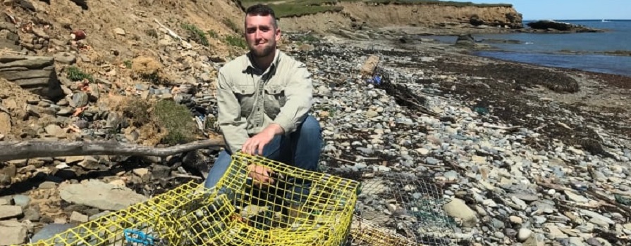 A man crouches on a rocky shore. In front of him is a broken lobster trap.