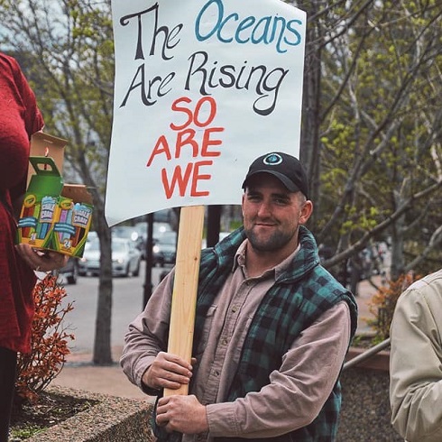 A smiling man wearing a hat holds a sign at a protest that reads, "The oceans are rising, so are we."