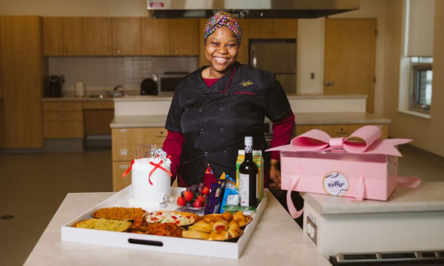 Nigerian woman in a chef's jacket smiles with an array of food and a cake in front of her.