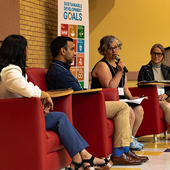 Four individuals sit on red chairs at the front of a recent conference. One individual is holding a microphone as she speaks to the crowd.