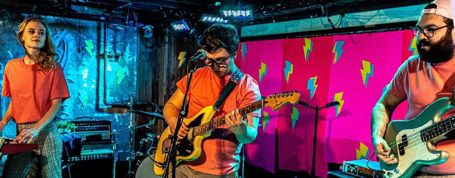 Two band members wearing orange shirts. The man is playing guitar and the woman is playing keyboards.