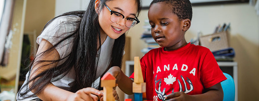 Amelia Dinh, an early childhood educator, smiles at a child in her care across a table as they play with wooden blocks.