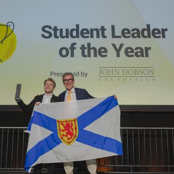 NSCC grad stands on stage holding a Nova Scotia flag, accepting the Enactus Student of the Year award.