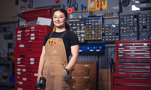 Hilary stands in a carpentry workshop. She wears a t-shirt, overalls, safety glasses and holds a drill in her right hand. Her left hand is in her pocket.