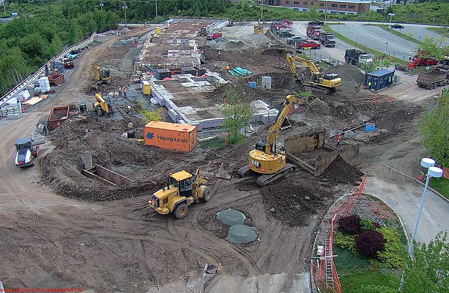 A drone flies over a construction site that is in the process of breaking ground near an empty parking lot. Tractors and trucks are pictured in the dirt