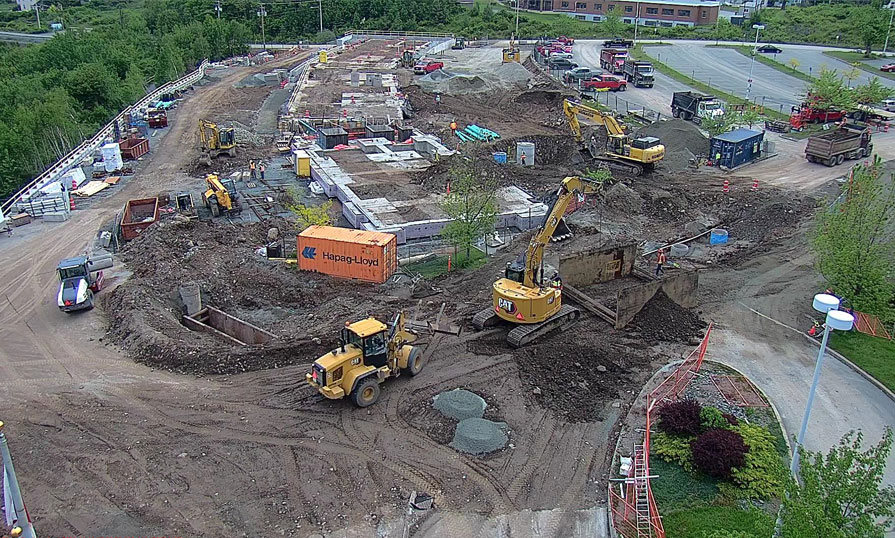 A drone flies over a construction site that is in the process of breaking ground near an empty parking lot. Tractors and trucks are pictured in the dirt