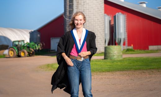 Image shows a female student wearing their graduation gown outside of a red barn.