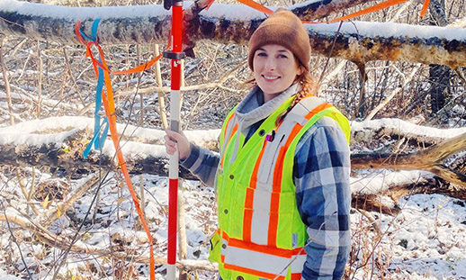 Kailey stands in a snowy wood wearing a safety west and holding surveying equipment.