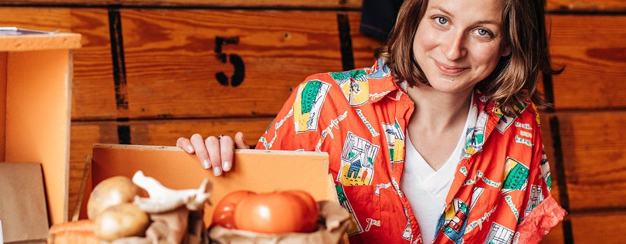 A woman in a patterned, orange shirt smiles at the camera. He hand is on a wooden crate containing produce.