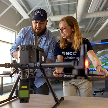 Two remote sensing students smile while learning to operate a drone that sits on a table in front of them 