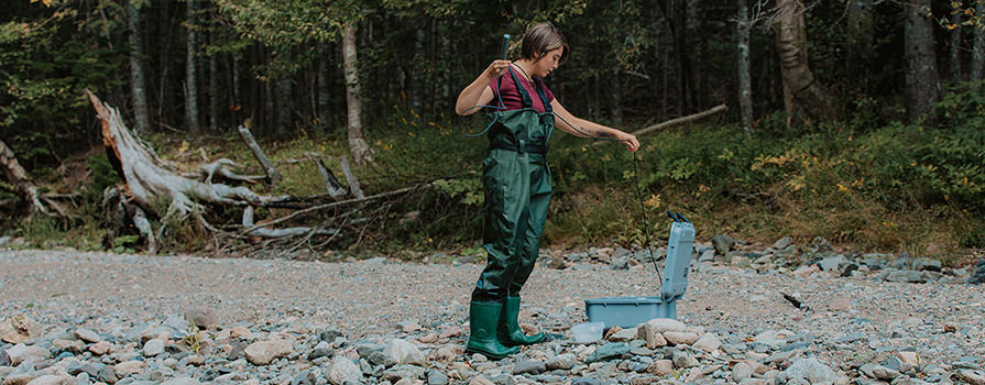 A woman in hip waders stands on a shoreline and takes a piece of equipment from a suitcase-style container.