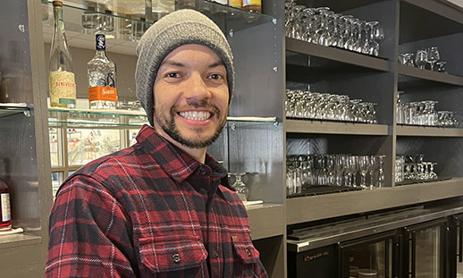 Leo wears a red plaid flannel shirt and stands in front of a well-stocked restaurant bar.