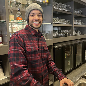 Leo wears a red plaid flannel shirt and stands in front of a well stocked restaurant bar.