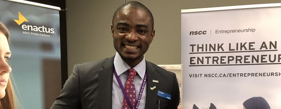 A man smiles broadly and looks at the camera. He is in a business suit and standing in front of two pull-up banners. One banner says Enactus and the other says NSCC Entrepreneurship.