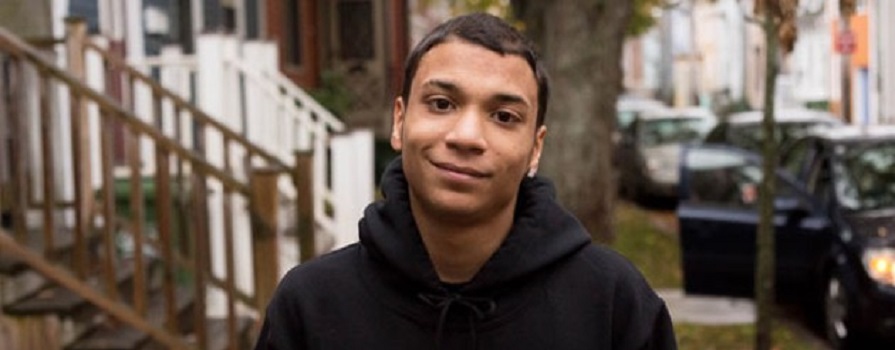 A head and shoulders shot of Unique Jones-MacKenzie. Unique is wearing a black hoodie and smiling slightly. A residential street can be seen behind him.