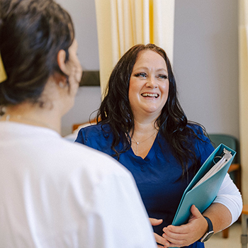 A person wearing blue scrubs, holding a binder while laughing with people wearing white lab coats look at them.