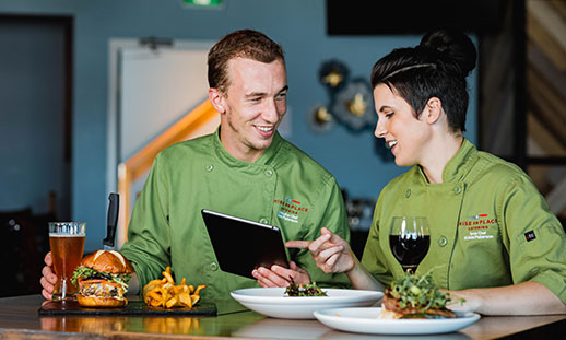 Chefs Mark Paterson and Krista LeTerte are shown. They are seated at a restaurant table and wearing green, chef’s jackets. They looking at an iPad, smiling and enjoying a meal and drinks.